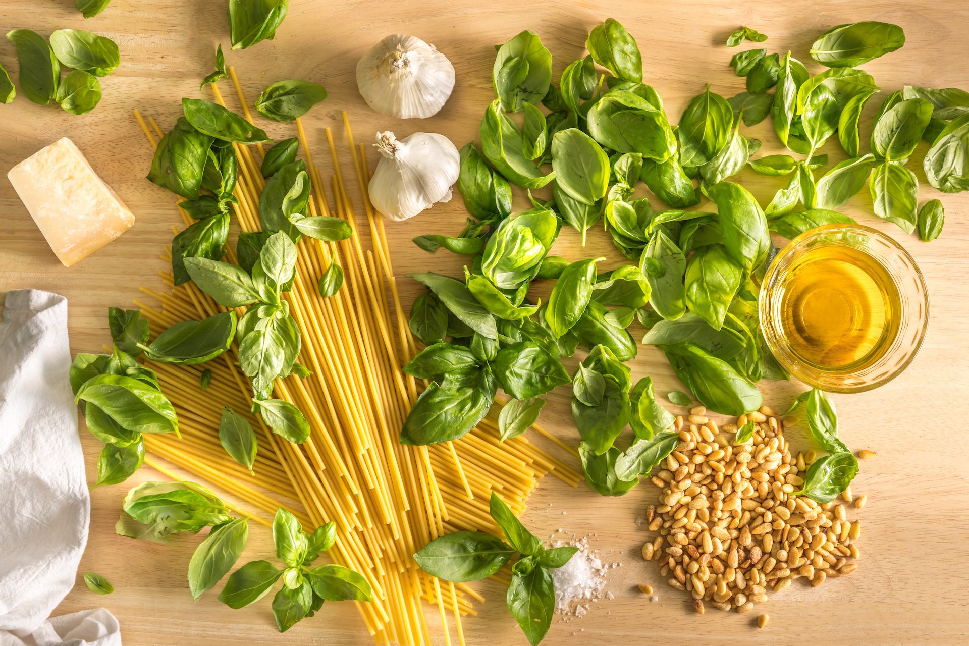 pasta, basil leaves, garlic, pine nuts, and olive oil arranged on a wooden counter