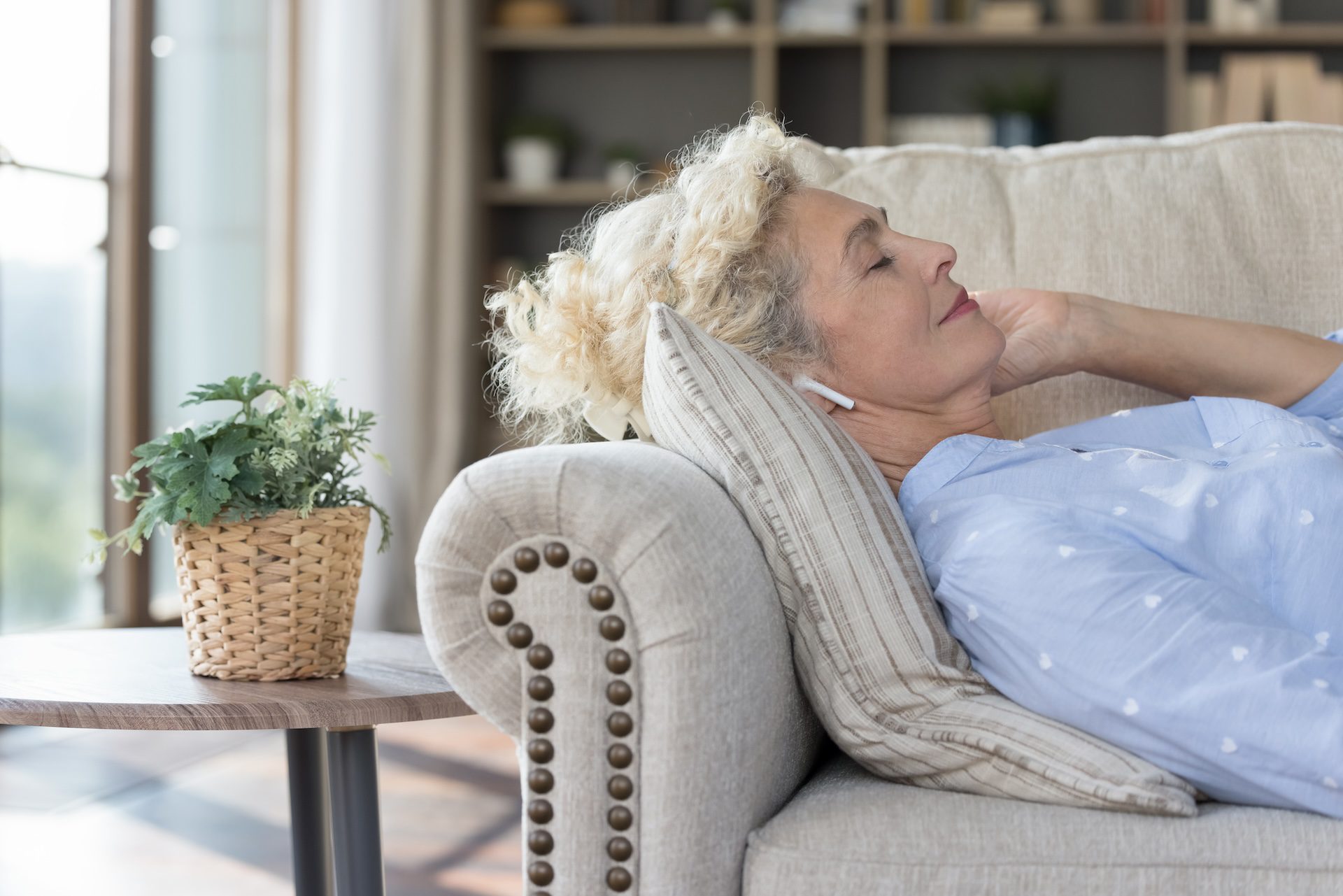 Caucasian mature woman listen music through modern wireless earphones, lying on cozy sofa in living room, chilling alone at home,