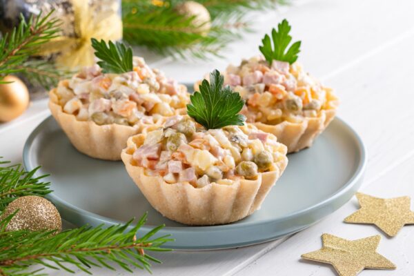 Olivier salad tartlets. Traditional russian appetizer for celebrating new year and christmas eve. White wooden table background. Fir tree branches, golden stars and balls. Copy space. Close up food.