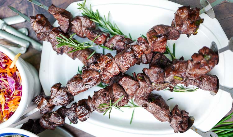 Skewers of grilled lamb garnished with rosemary on a white platter